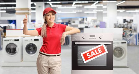 Photo for Female shop asisstant standing next to an electrcal oven inside a store and gesturing thumbs up - Royalty Free Image