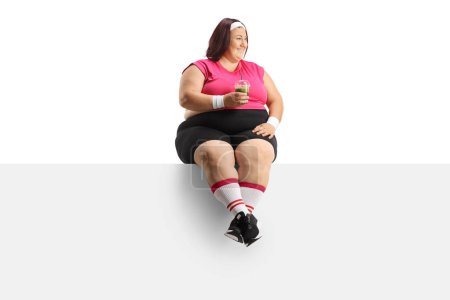Foto de Mujer con sobrepeso sonriente sosteniendo un batido verde saludable y sentado en un panel en blanco aislado sobre fondo blanco - Imagen libre de derechos