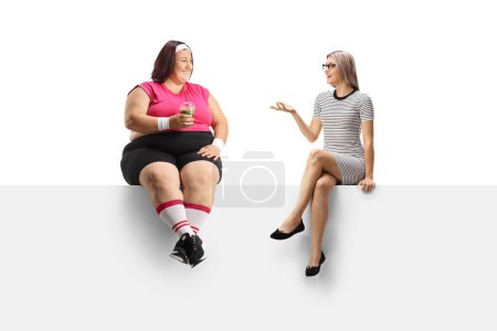 Téléchargez les photos : Femme mince parlant à une femme en surpoids tenant un smoothie vert sain et assis sur un panneau vierge isolé sur fond blanc - en image libre de droit