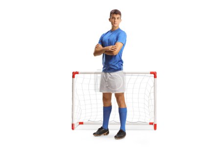Foto de Retrato completo de un joven futbolista en un jersey azul posando con los brazos cruzados frente a un mini gol aislado sobre fondo blanco - Imagen libre de derechos