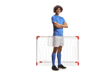 Foto de Retrato completo de un futbolista afroamericano en un jersey azul posando con los brazos cruzados frente a un mini gol aislado sobre fondo blanco - Imagen libre de derechos