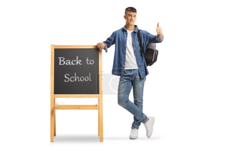 Photo for Full length portrait of a male teenage student with a backpack leaning on a blackboard with text back to school and gesturing thumbs up isolated on white background - Royalty Free Image