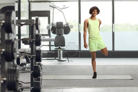 Téléchargez les photos : Portrait complet d'un jeune homme afro-américain en tenue de sport étirant la jambe au gymnase - en image libre de droit