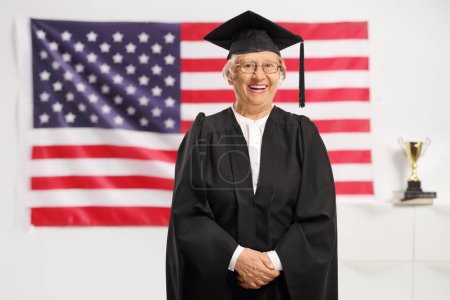 Photo for Elderly female wearing a graduation gown and smiling in front of a USA flag - Royalty Free Image