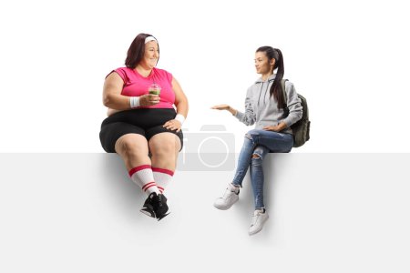 Foto de Estudiante hablando con una mujer con sobrepeso sosteniendo un batido verde saludable y sentada en un panel en blanco aislada sobre fondo blanco - Imagen libre de derechos