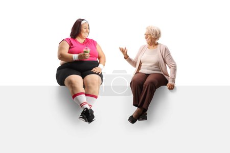 Téléchargez les photos : Femme âgée assise sur un panneau et parlant à une femme en surpoids tenant un smoothie vert sain isolé sur fond blanc - en image libre de droit