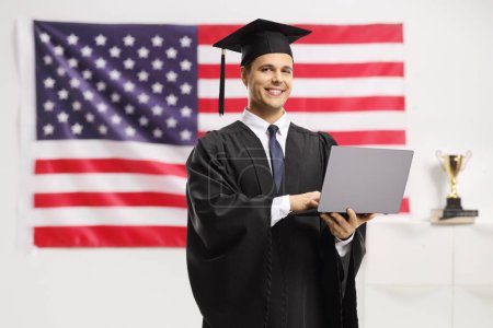 Foto de Hombre con un vestido de graduación negro y una gorra sosteniendo una computadora portátil frente a una bandera de EE.UU. - Imagen libre de derechos