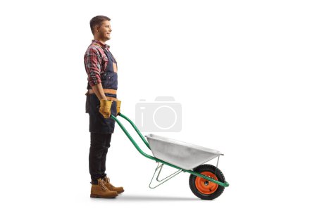 Photo for Full length profile shot of a farmer standing with an empty wheelbarrow solated on white background - Royalty Free Image