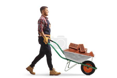 Photo for Full length profile shot of a young man pushing a wheelbarrow with bricks isolated on white background - Royalty Free Image