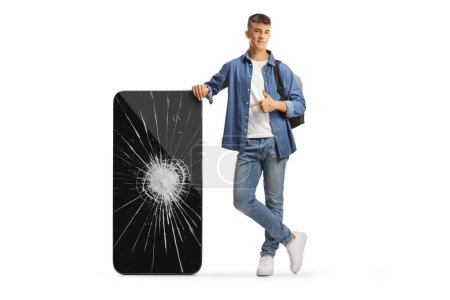 Photo for Male student with a backpack leaning on a big mobile phone and pointing at a broken screen isolated on white background - Royalty Free Image
