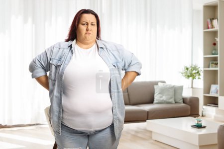 Foto de Mujer gruñona con sobrepeso de pie con las manos en la cintura en casa en una sala de estar - Imagen libre de derechos