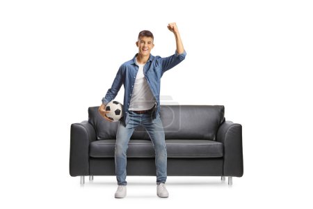Téléchargez les photos : Joyeux jeune homme avec un ballon de football acclamant devant un canapé isolé sur fond blanc - en image libre de droit