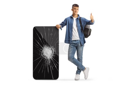 Photo for Male student with a backpack leaning on a broken mobile phone and gesturing thumbs up isolated on white background - Royalty Free Image