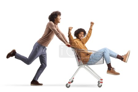 Photo for Young man pushing a happy african american young man inside a shopping cart isolated on white background - Royalty Free Image