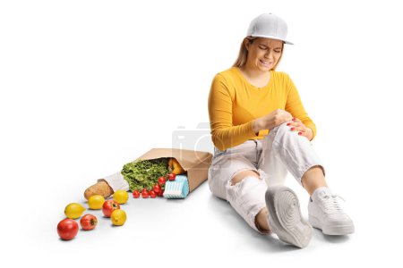 Photo for Young female with groceries sitting on the floor and holding her painful knee isolated on white background - Royalty Free Image