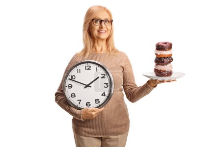 Foto de Mujer madura sosteniendo rosquillas en un plato y un reloj aislado sobre fondo blanco - Imagen libre de derechos