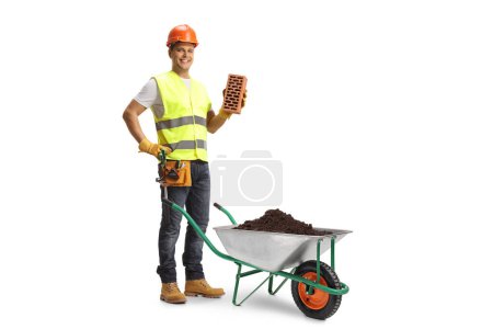 Téléchargez les photos : Ouvrier de la construction tenant une brique et debout derrière une brouette avec de la terre isolée sur fond blanc - en image libre de droit