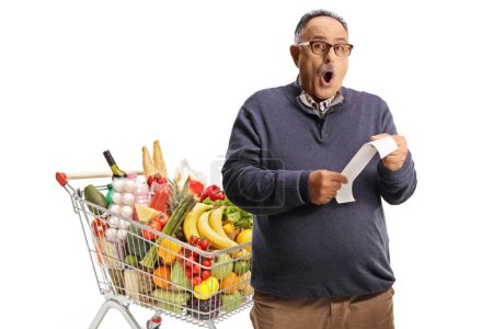 Photo for Shocked mature man with a shopping cart holding a bill isolated on white background - Royalty Free Image
