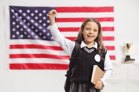 Foto de Feliz colegiala con una mochila y un libro haciendo gestos con la mano frente a una bandera americana - Imagen libre de derechos