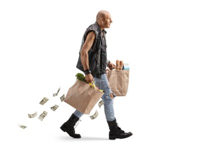 Photo for Full length profile shot of a punk man in a leather vest carrying groceries with money falling from the bags isolated on white background - Royalty Free Image