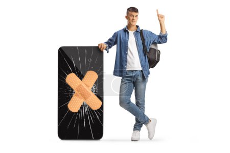 Foto de Estudiante masculino con una mochila apoyada en un teléfono móvil roto fijado con una venda y apuntando hacia arriba aislado sobre fondo blanco - Imagen libre de derechos