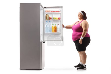 Téléchargez les photos : Femme corpulente debout devant un réfrigérateur ouvert et souriant isolé sur fond blanc - en image libre de droit