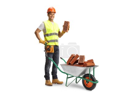 Photo for Construction worker holding a brick and standing behind a wheelbarrow with pile of bricks isolated on white background - Royalty Free Image