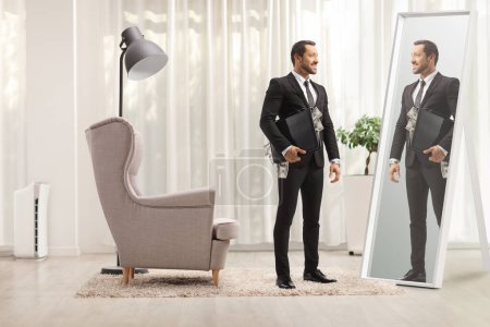 Téléchargez les photos : Homme d'affaires debout avec une mallette pleine d'argent à la maison et regardant dans un miroir - en image libre de droit