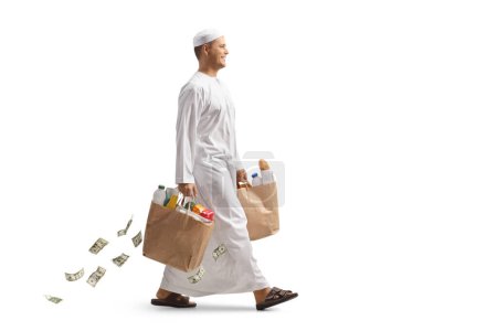Téléchargez les photos : Homme arabe en vêtements ethniques marchant et portant des sacs d'épicerie et de l'argent tombant derrière isolé sur fond blanc - en image libre de droit