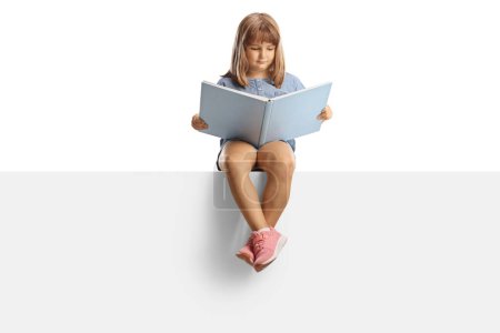 Photo for Little girl sitting on a blank panel and reading a book isolated on white background - Royalty Free Image