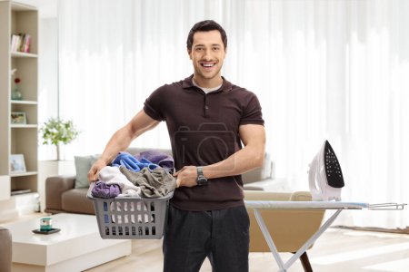 Photo for Guy holding a laundry basket full of clothes in front of an ironing board in a living room - Royalty Free Image