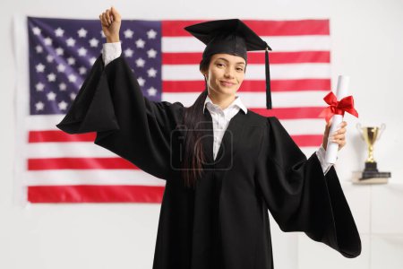 Téléchargez les photos : Joyeux srudent féminin dans une robe de remise des diplômes tenant un diplôme et geste avec la main devant un drapeau des États-Unis - en image libre de droit