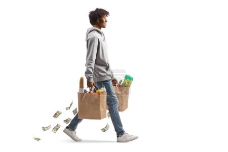 Foto de Tiro de perfil completo de un joven afroamericano con bolsas de comestibles caminando y perdiendo dinero aislado sobre fondo blanco - Imagen libre de derechos
