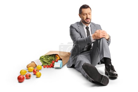 Téléchargez les photos : Homme d'affaires avec un sac d'épicerie assis sur le sol et tenant son genou douloureux isolé sur fond blanc - en image libre de droit