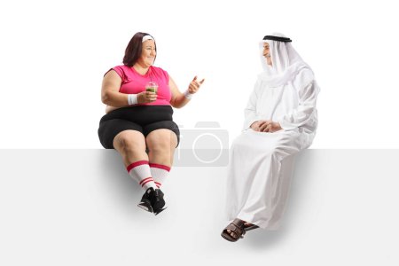 Téléchargez les photos : Femme corpulente en tenue de sport assise sur un panneau et parlant à un homme arabe mature isolé sur fond blanc - en image libre de droit