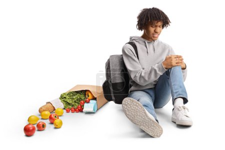 Téléchargez les photos : Étudiant afro-américain avec un sac d'épicerie assis sur le sol et tenant son genou blessé isolé sur fond blanc - en image libre de droit