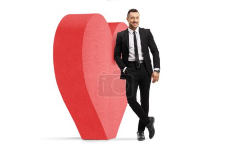 Photo for Full length portrait of a young man in a black suit and tie leaning on big red heart isolated on white background - Royalty Free Image