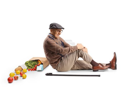 Foto de Hombre de edad avanzada con una bolsa de rocery sentado en el suelo y sosteniendo su pierna aislada sobre fondo blanco - Imagen libre de derechos