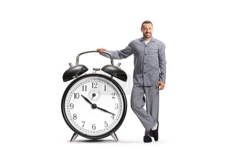 Foto de Hombre sonriente en pijama apoyado en un gran despertador aislado sobre fondo blanco - Imagen libre de derechos