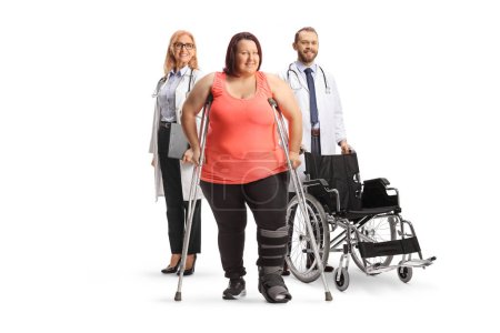 Photo for Corpulent woman wearing a foot brace and standing with crutches in front of a doctors with a wheelchair isolated on white background - Royalty Free Image
