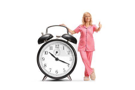 Photo for Woman in pajamas leaning on a big alarm clock and gesturing thumbs up isolated on white background - Royalty Free Image