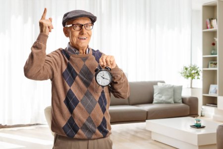 Foto de Anciano sosteniendo un despertador y señalando hacia arriba en su casa en una sala de estar - Imagen libre de derechos