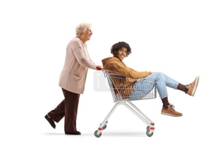 Photo for Elderly woman pushing an african american guy in a shopping cart isolated on white background - Royalty Free Image