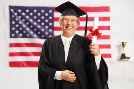 Foto de Mujer mayor en un vestido de graduado sosteniendo un título y sonriendo con la bandera de EE.UU. en el fondo - Imagen libre de derechos