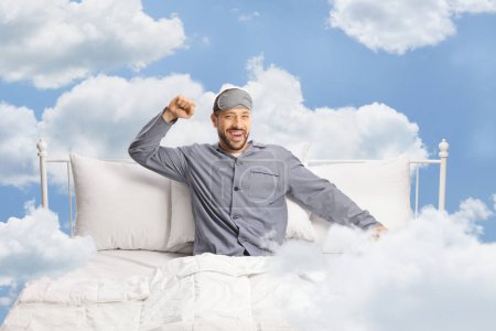 Téléchargez les photos : Heureux jeune homme avec un masque de sommeil qui s'étend dans un lit confortable dans les nuages - en image libre de droit