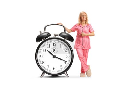 Téléchargez les photos : Belle femme mature en pyjama pointant vers un grand réveil isolé sur fond blanc - en image libre de droit