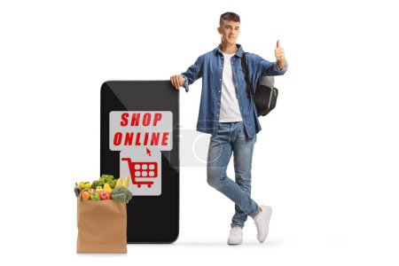 Téléchargez les photos : Homme étudiant geste pouces levés et penché sur un téléphone portable avec une application de magasinage en ligne et un sac d'épicerie isolé sur fond blanc - en image libre de droit