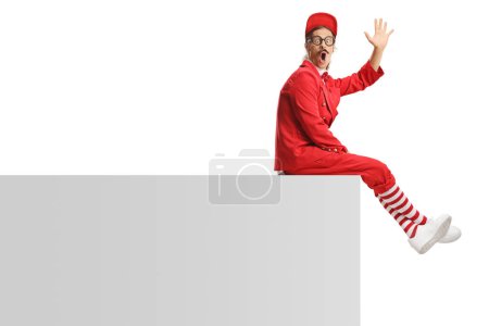 Téléchargez les photos : Animateur drôle en costume rouge assis sur un mur et agitant isolé sur fond blanc - en image libre de droit