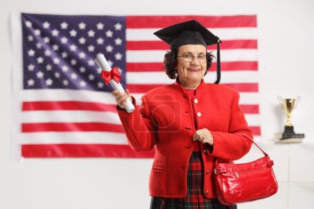 Téléchargez les photos : Dame âgée avec un diplôme et un chapeau de graduation souriant à la caméra devant un drapeau des États-Unis - en image libre de droit