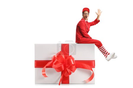 Téléchargez les photos : Animateur en costume rouge assis sur une grande boîte cadeau et agitant isolé sur fond blanc - en image libre de droit